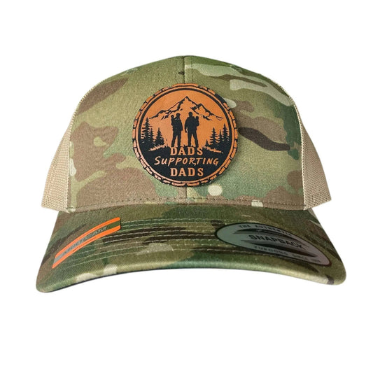 A camo leather patch trucker hat that says "Dads Supporting Dads" on the patch with the silhouettes of 2 men with children and mountains with beige mesh on the back half of the hat on a white background.