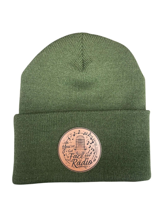 A green leather patch beanie engraved with "You've Got A Face For Radio" wording overlaid with musical notes and a microphone, all set on a white background.