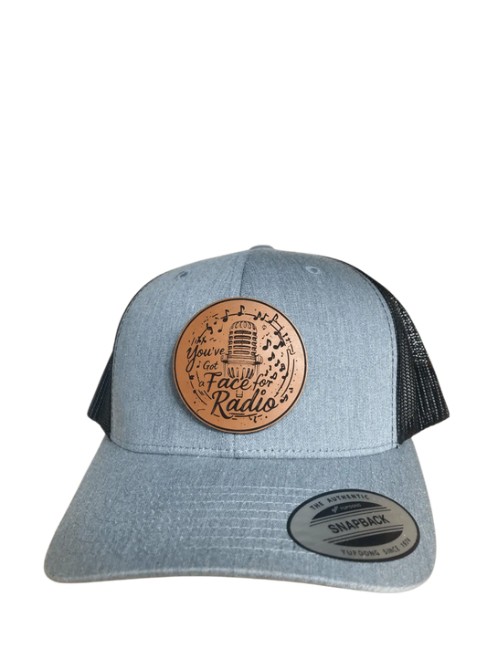 A grey leather patch trucker hat that says "You've Got A Face For Radio" on the patch with musical notes and a microphone with black mesh on the back half of the hat on a transparent background.