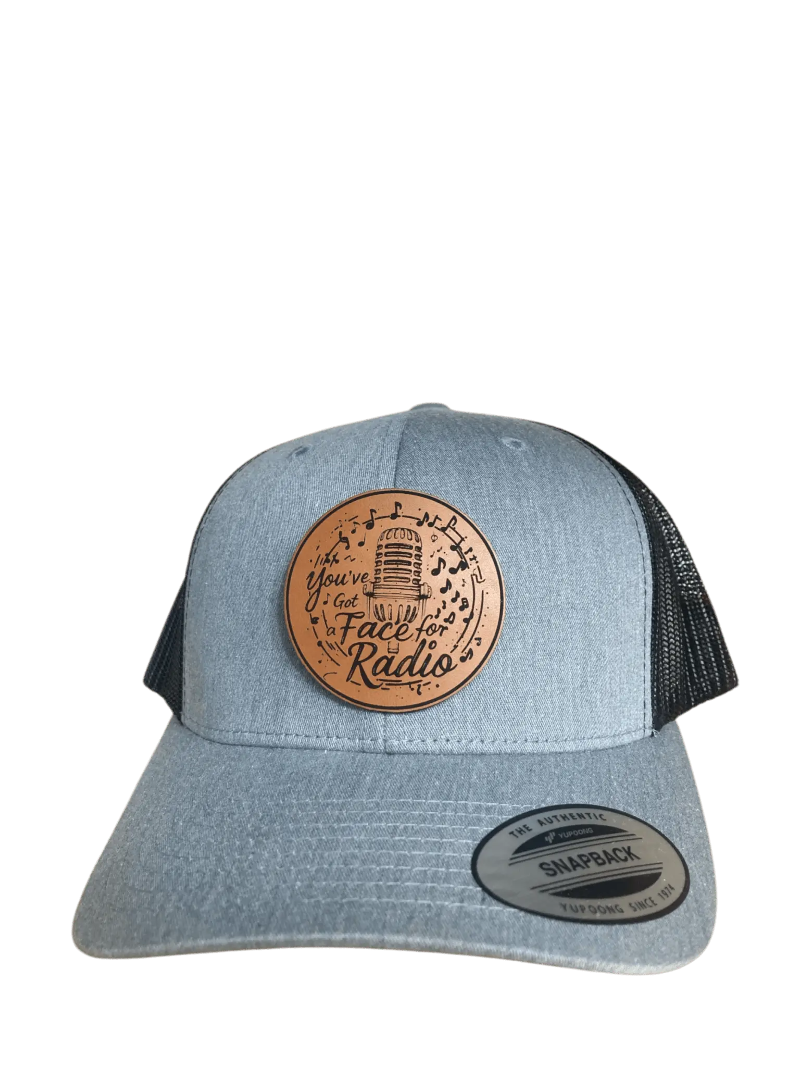 A grey leather patch trucker hat that says "You've Got A Face For Radio" on the patch with musical notes and a microphone with black mesh on the back half of the hat on a transparent background.