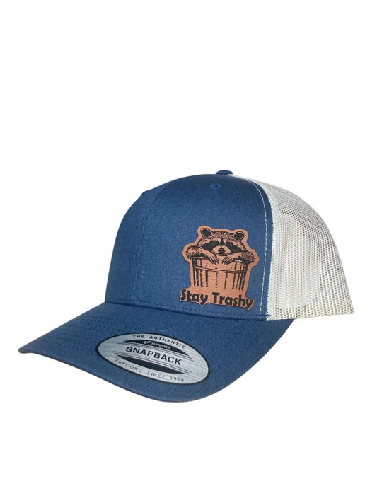 A blue leather patch trucker hat that says "Stay Trashy" on the patch with the picture of a raccoon peaking out of a trash can with white mesh on the back of the hat on a white background.