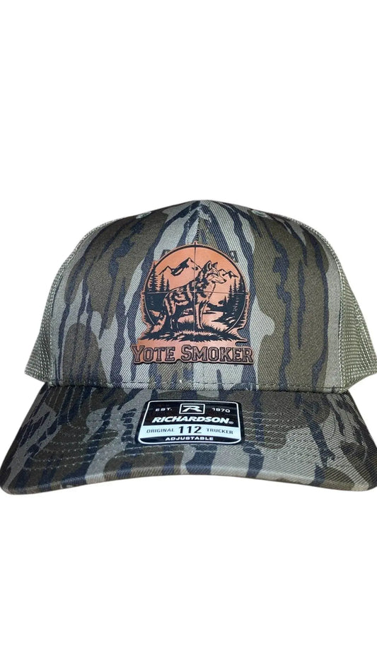 A green camo leather patch trucker hat that says "Yote Smoker" on the patch with a pitcher of a coyote and mountains with green mesh on the back half of the hat on a white background.
