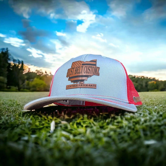 A grey custom engraved leather patch trucker hat engraved with custom business branding with orange mesh on the back half of the hat sitting on a grass with a blue sky background.