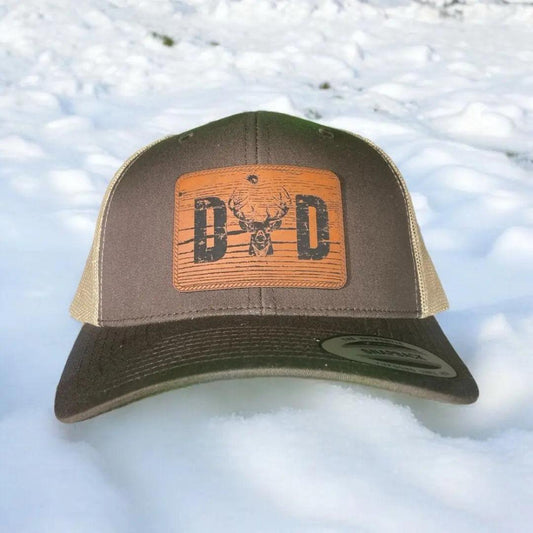 A brown leather patch trucker hat that says "Dad" on the patch with a whitetail buck replacing the A with beige mesh on the back half of the hat on a snowy background.