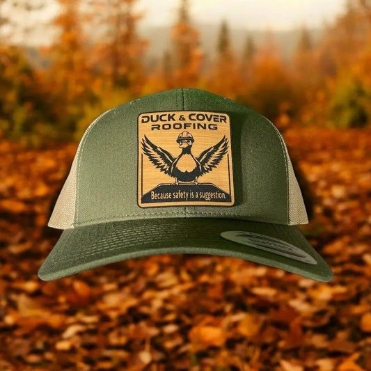 A green leather patch trucker hat that says "Duck & Cover Roofing - Because safety is a suggestion" on the patch with a picture of a duck wearing a hard hat with beige mesh on the back half of the hat on a background of fall leaves.
