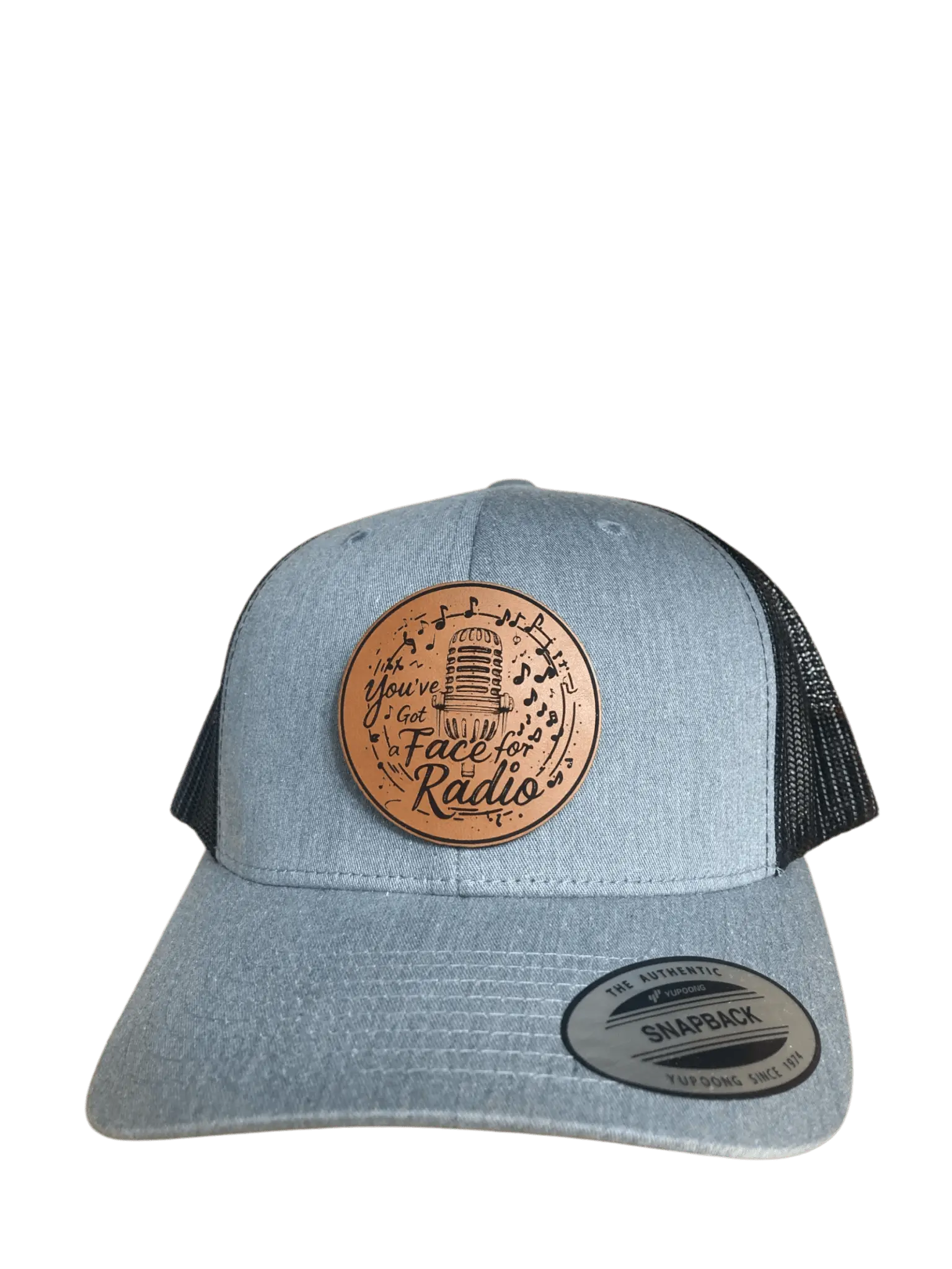 A grey leather patch trucker hat that says "You've Got A Face For Radio" on the patch with musical notes and a microphone with black mesh on the back half of the hat on a transparent background.