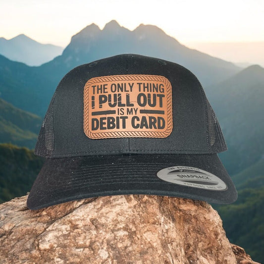 A black leather patch trucker hat with "The Only Thing I Pull Out Is My Debit Card" on the leather patch with black mesh on the back half with a mountainous background.