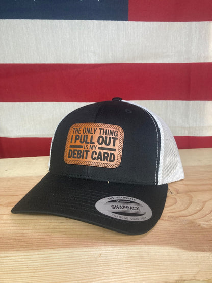 A black leather patch trucker hat with "The Only Thing I Pull Out Is My Debit Card" on the leather patch with white mesh on the back half with an American flag background.
