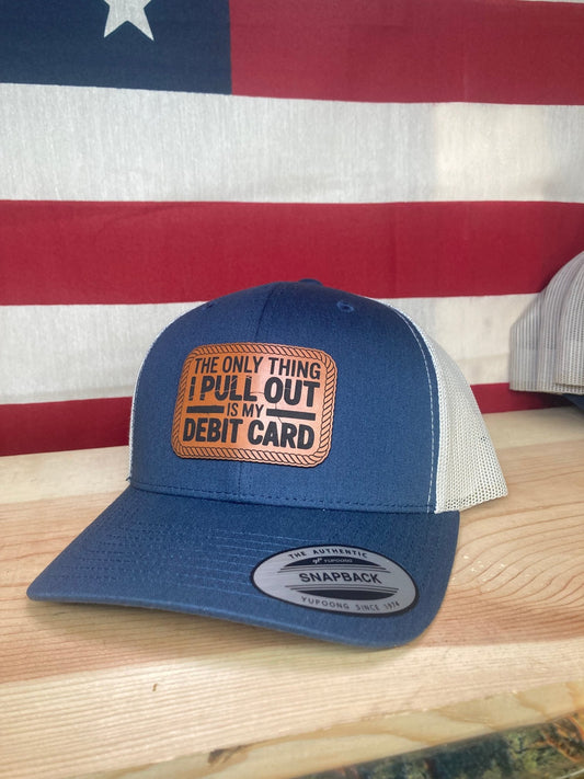 A blue leather patch trucker hat with "The Only Thing I Pull Out Is My Debit Card" on the leather patch with white mesh on the back half with an American flag background.