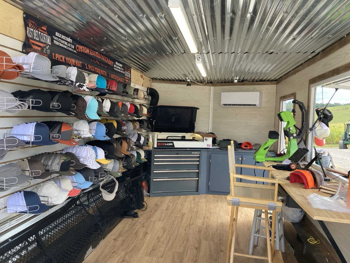 Inside the Rust Belt Customz trailer with a wall of hats, laser, and hat presses