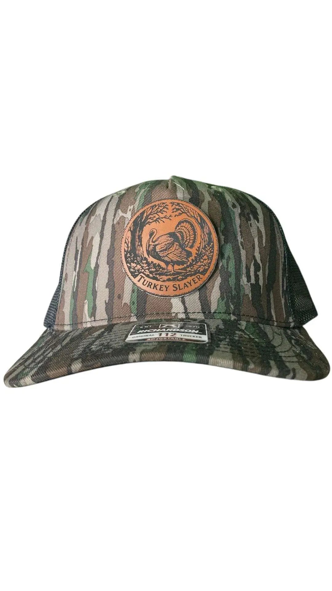 A camo leather patch trucker hat that says "Turkey Slayer" on the patch with a picture of a turkey with black mesh on the back half of the hat on a white background.