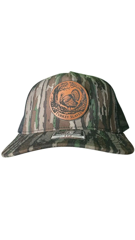 A camo leather patch trucker hat that says "Turkey Slayer" on the patch with a picture of a turkey with black mesh on the back half of the hat on a white background.